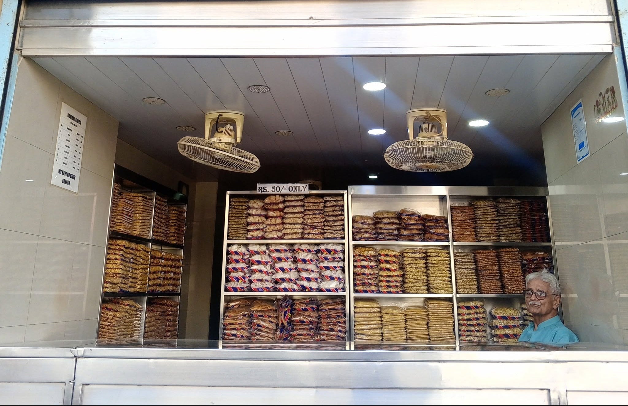 A storefront sign reading 'BOMBAY NIMCO We have no other branch' above a roll-up shutter, with shelves of packaged goods visible inside and a person partially visible at the lower right.